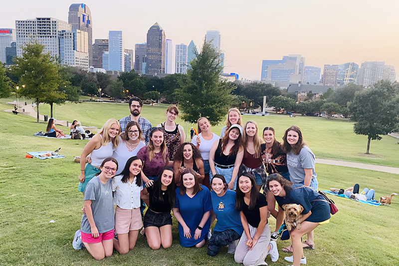 Students from the first, second, and third year class pose for a group photo at the park with a view of the Dallas skyline at dusk in the background.